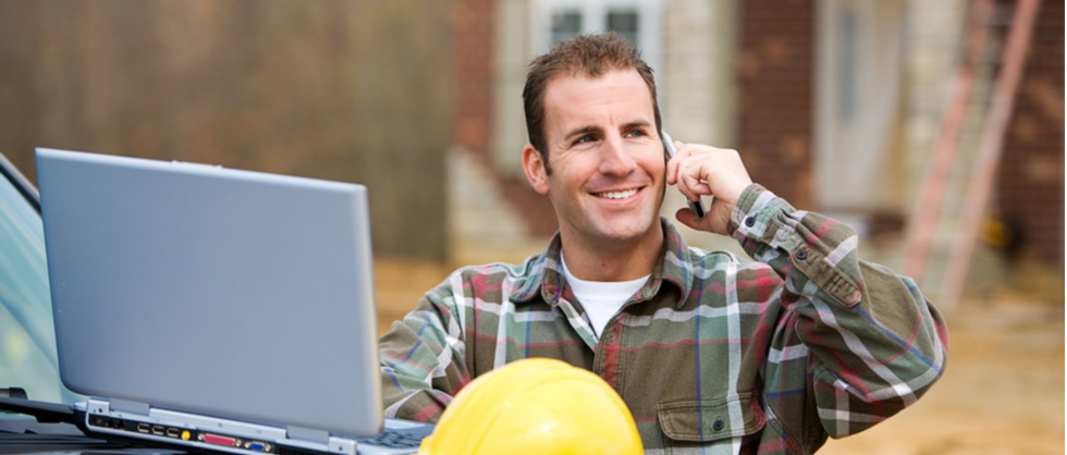 smiling man on a construction site with laptop open on his hood next to a yellow hard hat using mobile phone