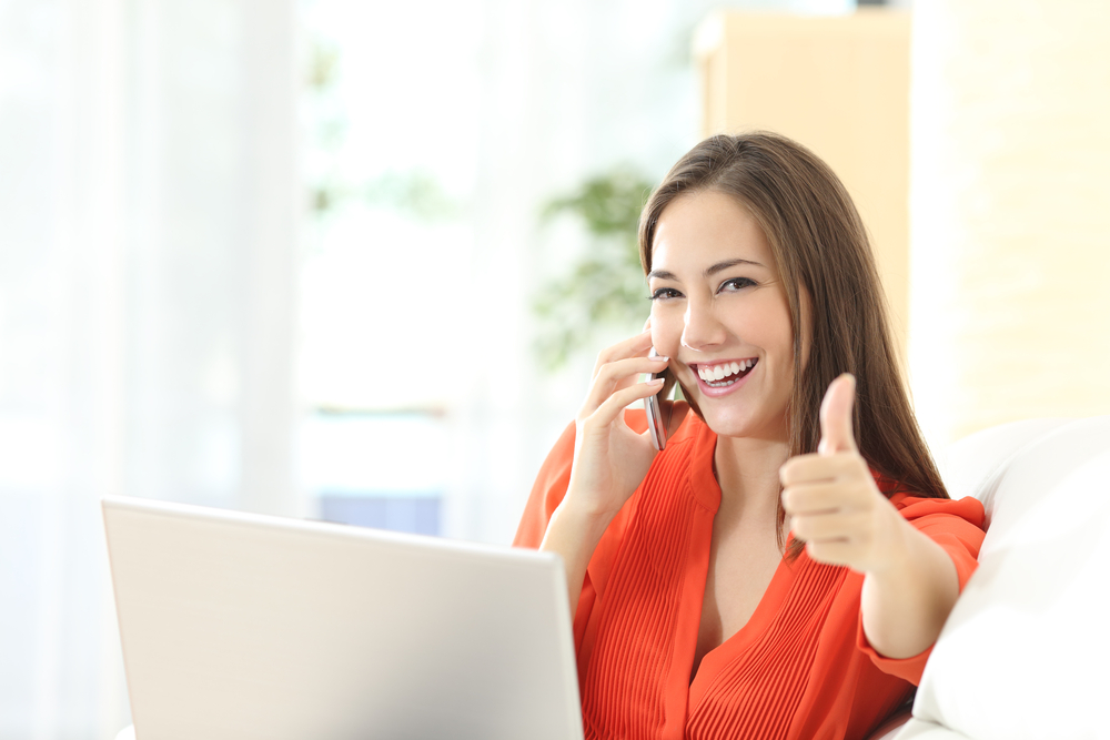 Happy young woman wearing a bright orange top on the phone in front of her computer showing a thumbs up