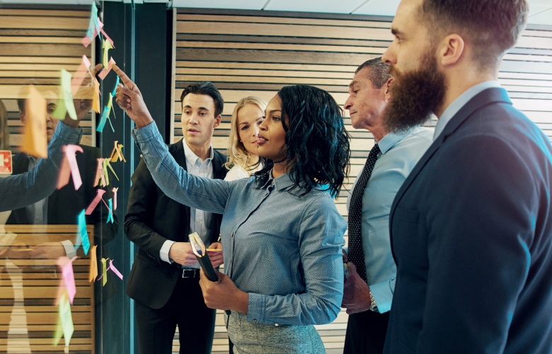 Group of business people standing in front of a glass wall covered in post it notes and a woman speaking as she points to something on the wall